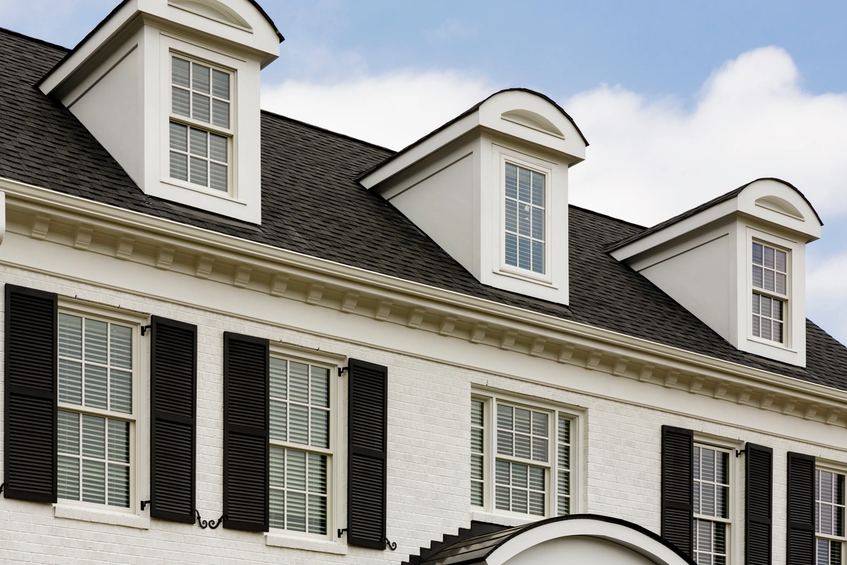 A close-up of the upper part of a white brick house with black shutters, three dormer windows on the roof, and a blue sky with clouds in the background, showcasing classic roofing Mount Vernon style.