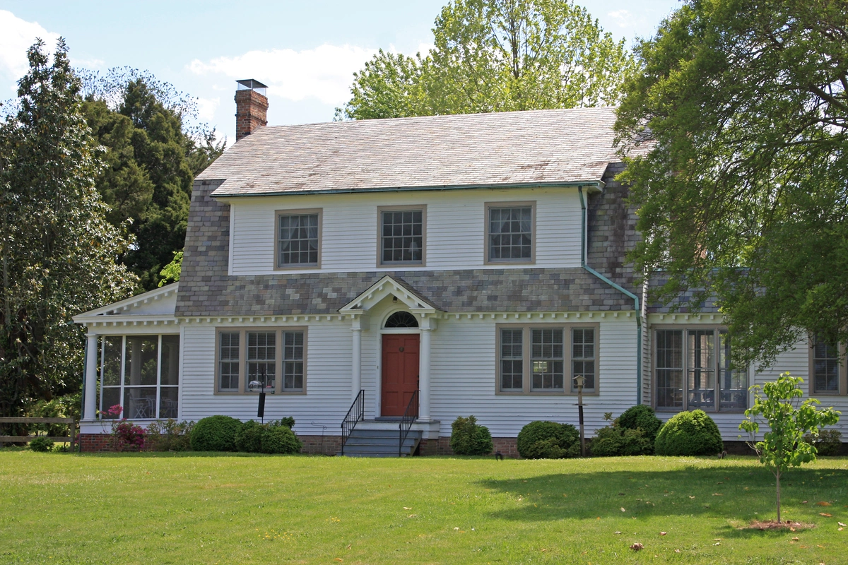 A two-story white house with a gray shingle roof, red front door, and multiple windows sits surrounded by green grass, bushes, and trees—showcasing expert roofing Falls Church style on a sunny day.