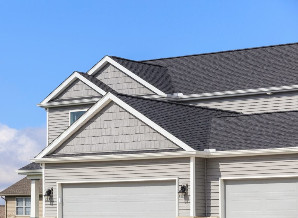 A close-up view of the upper exterior of a modern suburban house in McLean highlights quality roofing materials, light gray siding, white trim, asphalt shingle roofing, and double garage doors under a clear blue sky. Roofing Materials McLean
