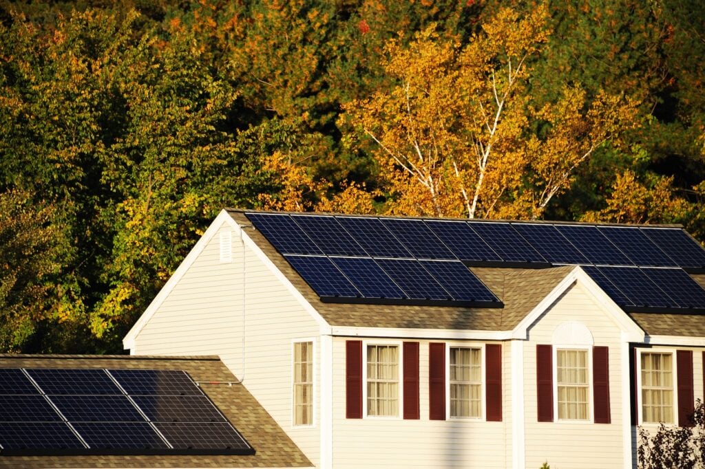 A house with dark solar panels on its roof is shown in front of trees with green and yellow autumn leaves, with sunlight illuminating the scene. Roof Maintenance McLean