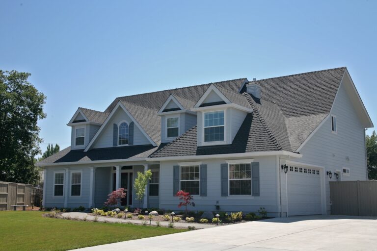 A large, two-story suburban house with gray siding, white trim, multiple gables, and a well-maintained front yard with shrubs and small trees under a clear blue sky.