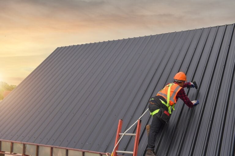 A construction worker in a safety harness, helmet, and reflective vest climbs a ladder onto a sloped metal roof at sunset, inspecting or working on the roofing panels.