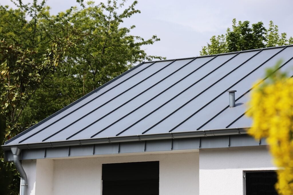 A modern house with a gray metal roof and white walls, showcasing premium roofing materials McLean, surrounded by green trees and a blurred yellow plant in the foreground. Roofing Materials McLean