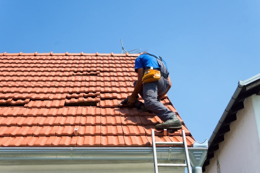 A worker on a ladder repairs red roof tiles on a house under a clear blue sky, wearing a blue shirt, gray pants, and a yellow tool belt—showcasing expert service and giving insight into Roof Repair Cost McLean.