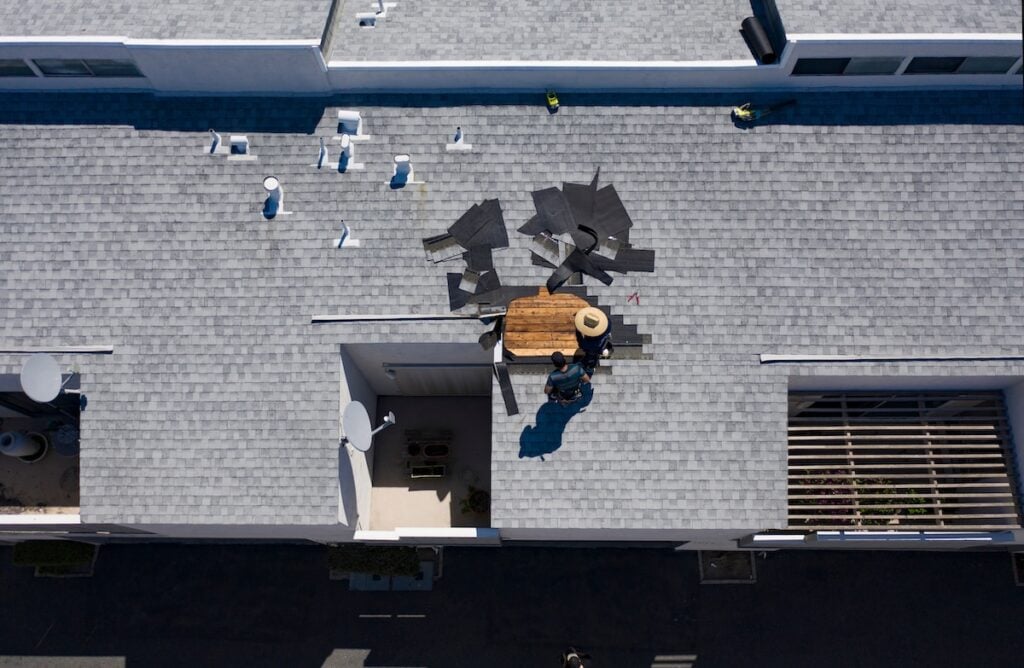 Aerial view of two workers repairing shingles on a gray shingled roof in daylight. Tools and shingles are scattered around their workspace. The building has a rooftop vent and a partially covered  patio below. McLean Roofing Companies