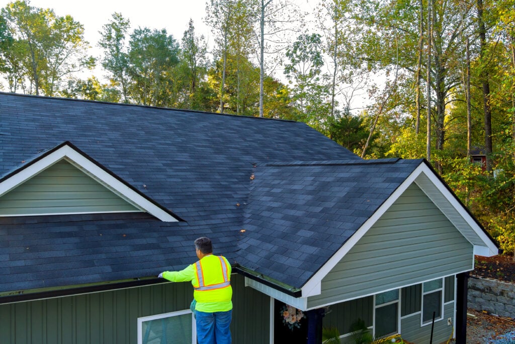 A person in a neon safety vest stands on a ladder, cleaning leaves from the gutter of a house with a dark gray shingled roof, surrounded by green trees.