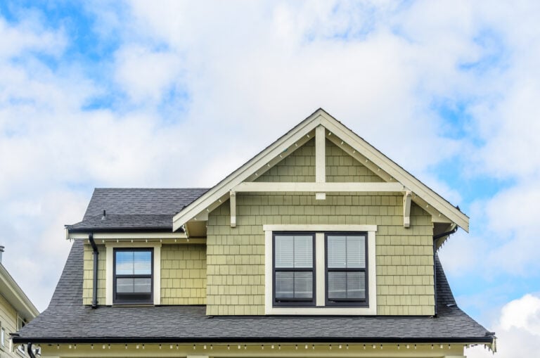 The upper facade of a house with light green siding, white trim, two windows with shutters, and a steep, dark gray roof hints at the style and potential Roof Repair Cost McLean homeowners might consider under a partly cloudy blue sky.