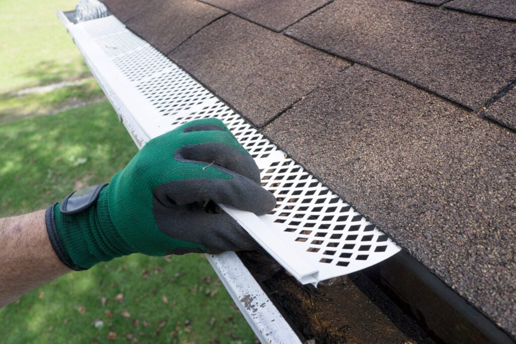 A person wearing a green work glove installs a white mesh gutter guard along the edge of a roof with brown shingles during a Roof Inspection McLean, placing it over an open gutter to prevent debris from entering.