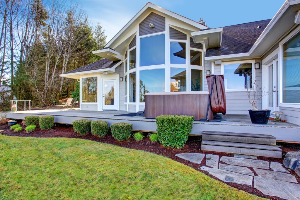Modern house with large windows, a wooden deck, hot tub, and trimmed bushes on a well-kept lawn, surrounded by tall trees under a clear blue sky—perfect for your Roof Inspection McLean needs. Roof Inspection McLean