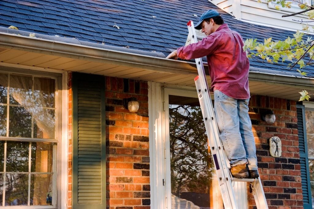 A person wearing a red shirt and blue jeans stands on a ladder, cleaning leaves from the gutter of a brick house with green shutters and a shingled roof.