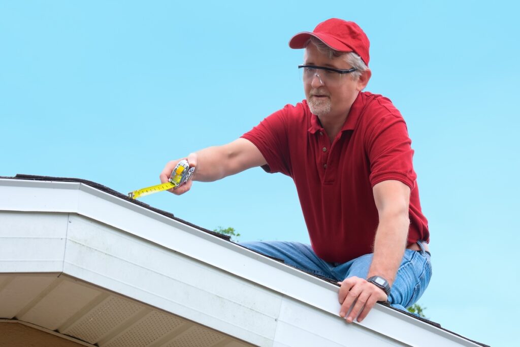 A man in a red shirt, cap, and safety glasses kneels on a roof, using a tape measure to check the edge—a perfect scene for your Roof Inspection Schedule in McLean. The sky is clear and blue in the background.