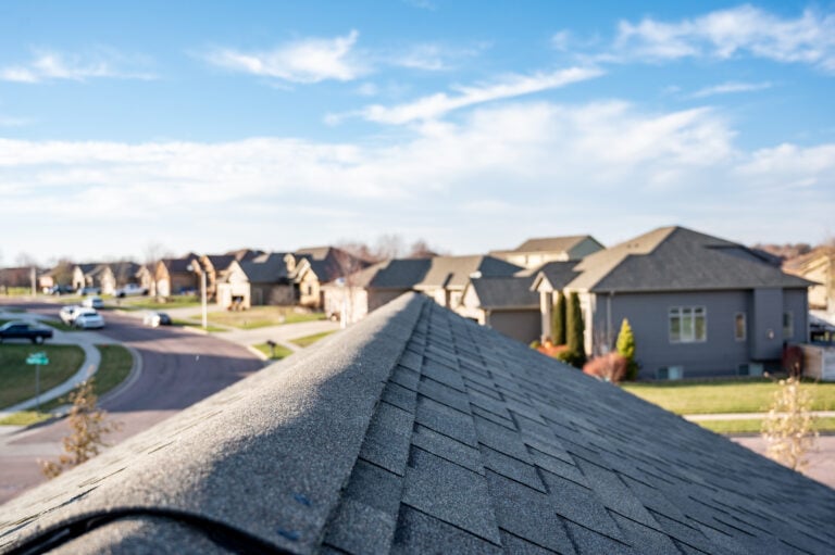 View from the peak of a shingled roof overlooking a suburban neighborhood with houses and lawns—an ideal vantage for a Roof Inspection Schedule McLean, under a partly cloudy blue sky and along a winding street.