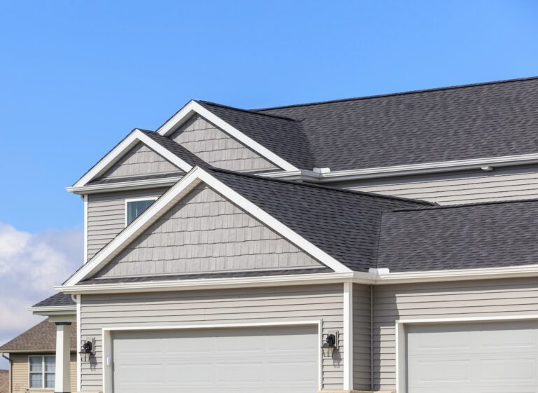 The image shows the upper portion of a modern suburban house with gray siding, white trim, three gabled roofs, and two closed white garage doors against a clear blue sky—an inspiring sight for anyone considering DIY Roof Maintenance McLean.