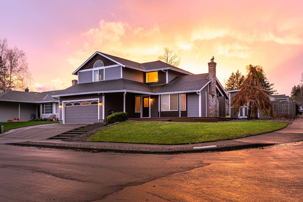 Two-story suburban house with a gray exterior, attached garage, and manicured lawn on a corner lot. The sky is vibrant with orange and pink hues from the sunset. The street and driveway appear wet.
