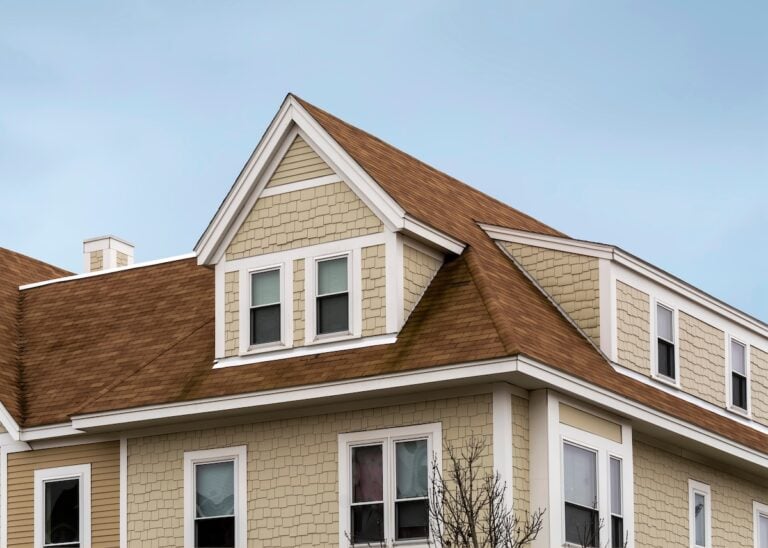 A beige house with brown shingles, white trim, and multiple windows sits under a blue sky. The new roof with steep gables and dormer windows enhances its classic, suburban appearance and adds significant home value in McLean.