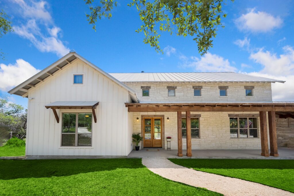 Roofing Trends McLean A modern farmhouse with white siding and stone exterior, wooden posts supporting a covered porch, large windows, and a neatly manicured lawn under a bright blue sky with scattered clouds.