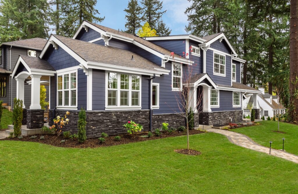 New Roof Home Value McLean A modern two-story house with blue siding, white trim, and stone accents in McLean. A new roof adds home value to the well-manicured lawn, small garden beds, and tall trees. A curved walkway leads to the front entrance.
