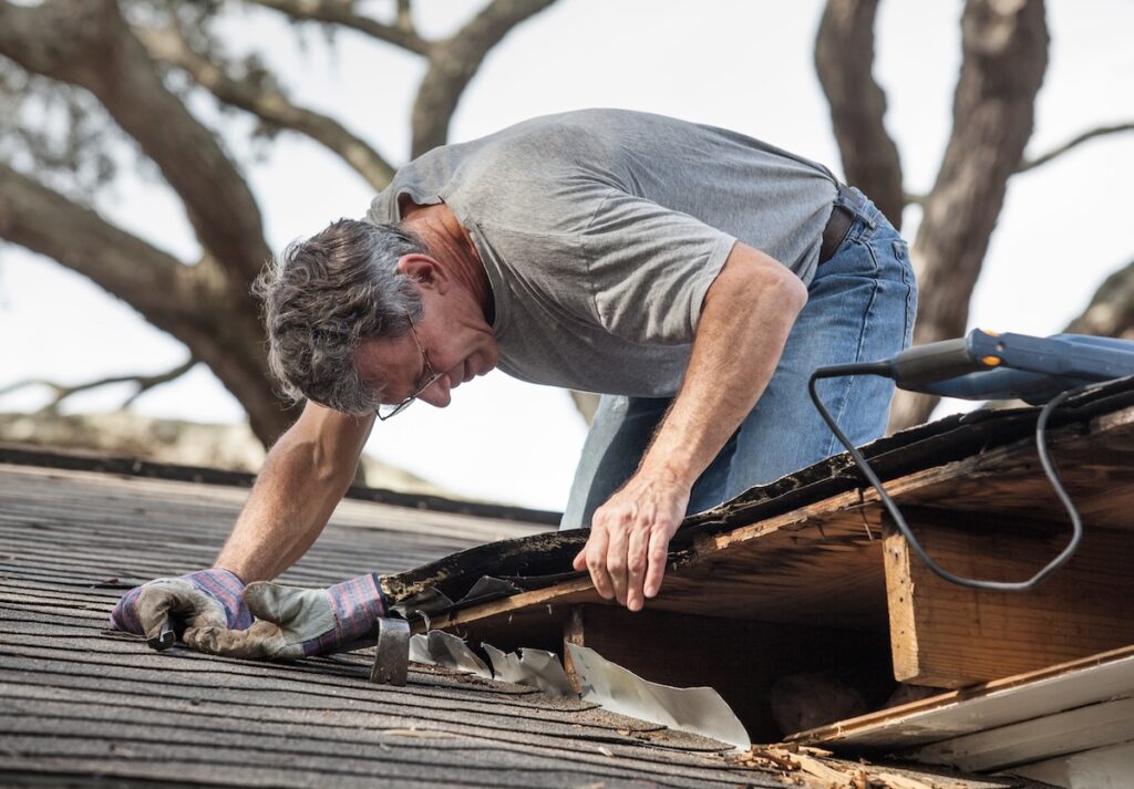 A man wearing glasses and gloves repairs the edge of a roof, carefully inspecting and lifting damaged shingles and wood during DIY Roof Maintenance in McLean. A power tool rests nearby, with large tree branches seen in the background. DIY Roof Maintenance McLean