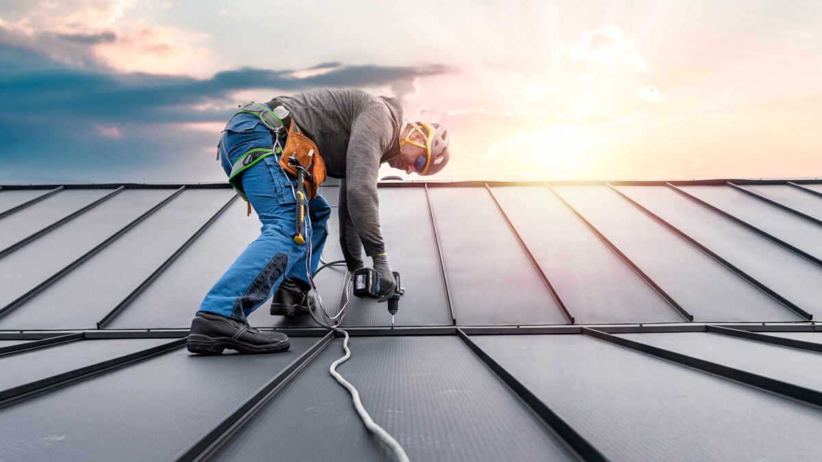 Roofing Company McLean A construction worker in safety gear uses a power tool on a metal roof at sunset, wearing a harness attached to a safety line for protection.