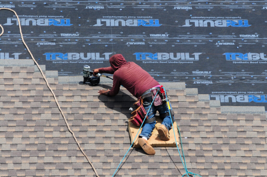 Roofing Company McLean A roofer wearing a red hoodie and safety gear is kneeling on a shingled roof, using a nail gun to install shingles with roofing underlayment visible above. Safety ropes are attached to the worker.