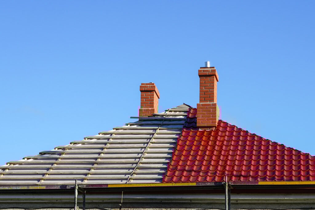Roofing Warranties McLean A house roof under construction, with the left side showing exposed wooden beams and the right side covered with red tiles. Two red brick chimneys rise from the roof against a clear blue sky.