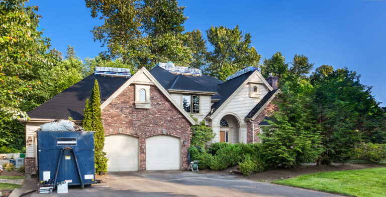Roofing Warranties McLean A brick house with two garages and tall trees in the background. Roofing materials are stacked on the roof, and a dark blue trailer is parked in the driveway, suggesting ongoing roof construction or repair work.