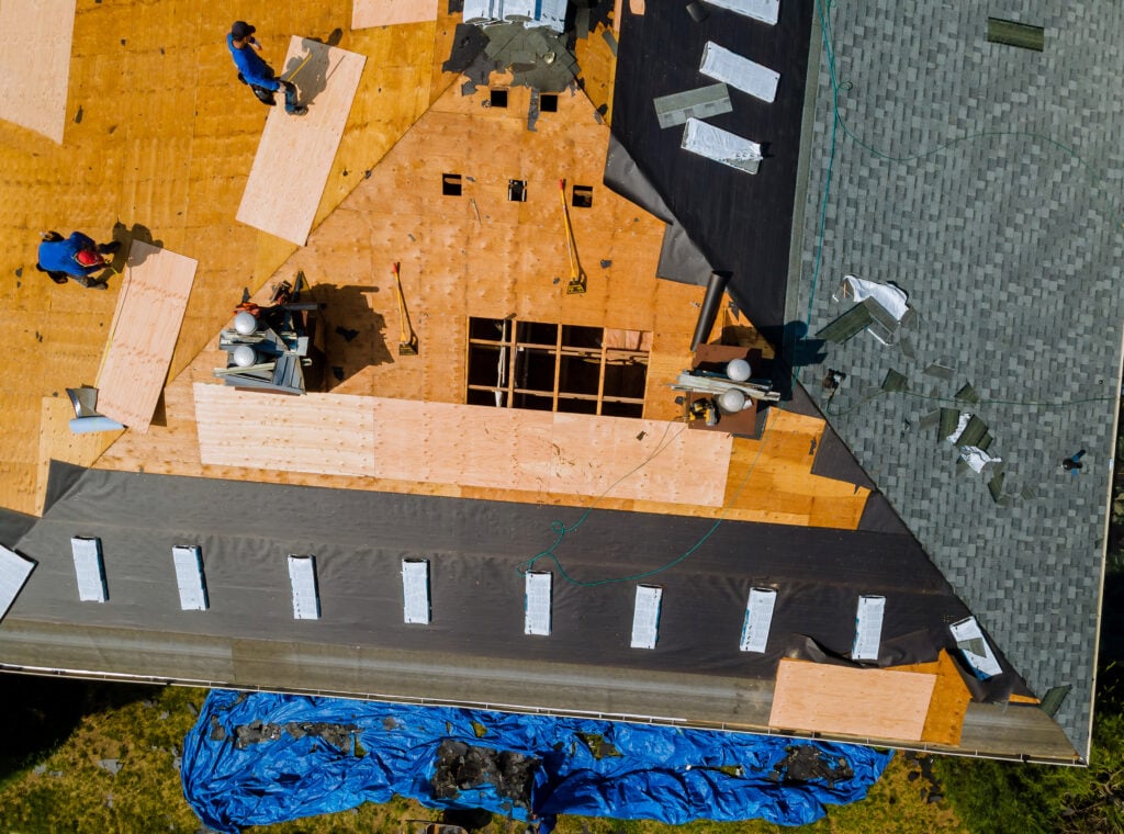 Aerial view of workers installing plywood sheets and roofing materials on a house roof in McLean, illustrating a typical roof replacement timeline. Some areas are shingled, others exposed, with tools and materials scattered and a blue tarp on the ground. Roof Replacement Timeline McLean