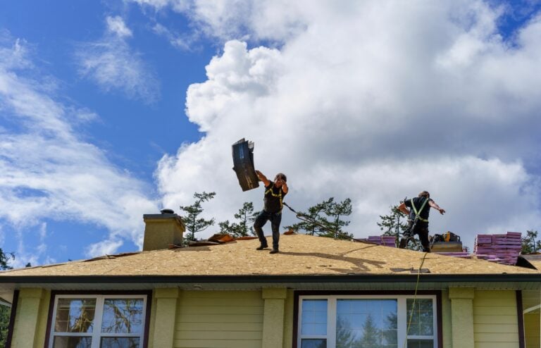 Two workers stand on the roof of a house under a blue sky with clouds. During this Roof Replacement McLean project, one lifts roofing materials while the other moves near stacked supplies, preparing the stripped roof for new shingles.