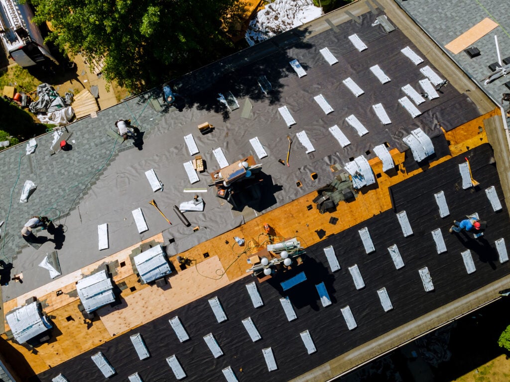 Aerial view of workers installing a new roof on a building during a Roof Replacement McLean project. Strips of roofing materials, tools, and bundles of shingles are scattered across the partially covered wooden roof surface. Roof Replacement McLean