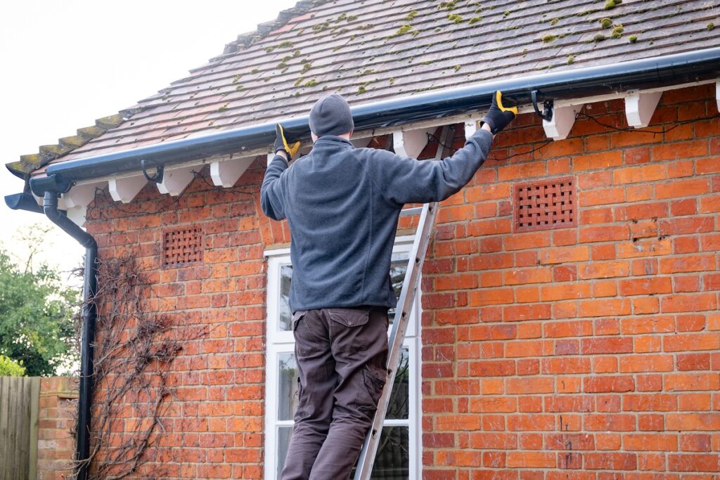 A person standing on a ladder is performing DIY roof maintenance in McLean, cleaning or inspecting the gutters on a brick house. The individual is wearing a gray jacket, brown pants, gloves, and a beanie.