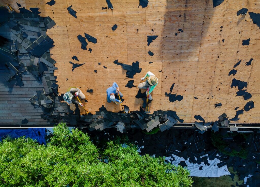 Roofing Warranties McLean Aerial view of three people removing old shingles from a rooftop, surrounded by debris. The roof is partially stripped, revealing wooden boards beneath, and greenery is visible at the bottom of the image.