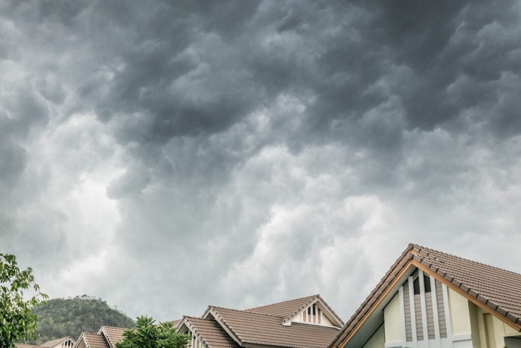 Dark storm clouds gather above a row of modern houses with brown tiled roofs, hinting at weather that may soon call for roof replacement McLean residents can trust. Trees line the foreground, with a forested hill looming in the background.