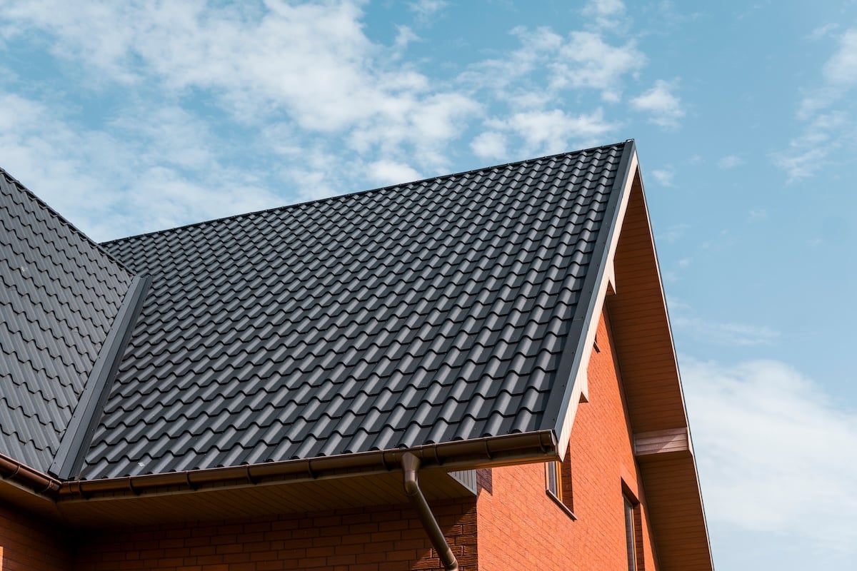 A close-up view of a house with a sloped black tiled roof and orange brick walls, set against a blue sky with scattered clouds.