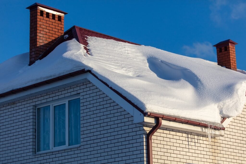 winter roof replacement Virginia A white brick house with two red brick chimneys, topped with a thick layer of snow on the roof. Icicles hang from the eaves, and a clear blue sky is visible in the background.