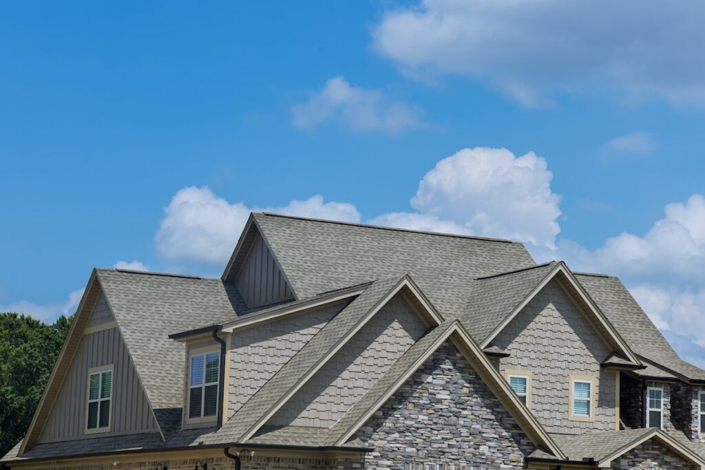 A close-up view of a house roof with multiple gables, gray shingles, and stone accents on the exterior, set against a blue sky—highlighting roofing warranties McLean homeowners can rely on.