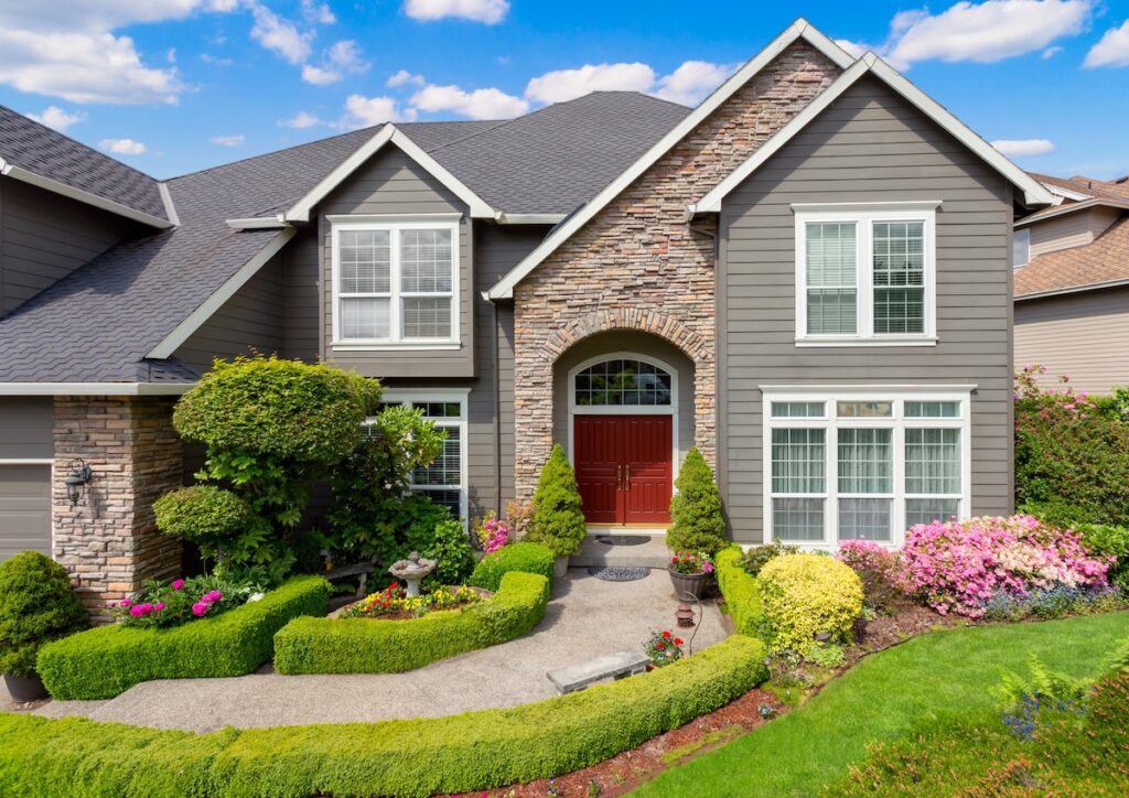 A modern suburban house with gray siding, stone accents, and red double front doors stands under a blue sky in McLean, featuring white-trimmed windows, a lush garden, and the peace of mind that comes with reliable roofing warranties.