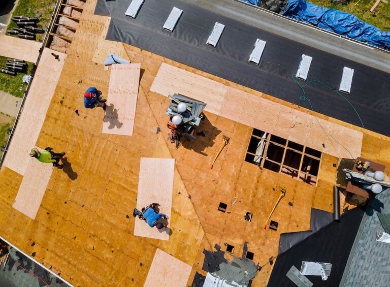Aerial view of workers installing plywood sheets on a house roof under construction, with tools and materials scattered around. Four people are visible working on different sections of the large rooftop.