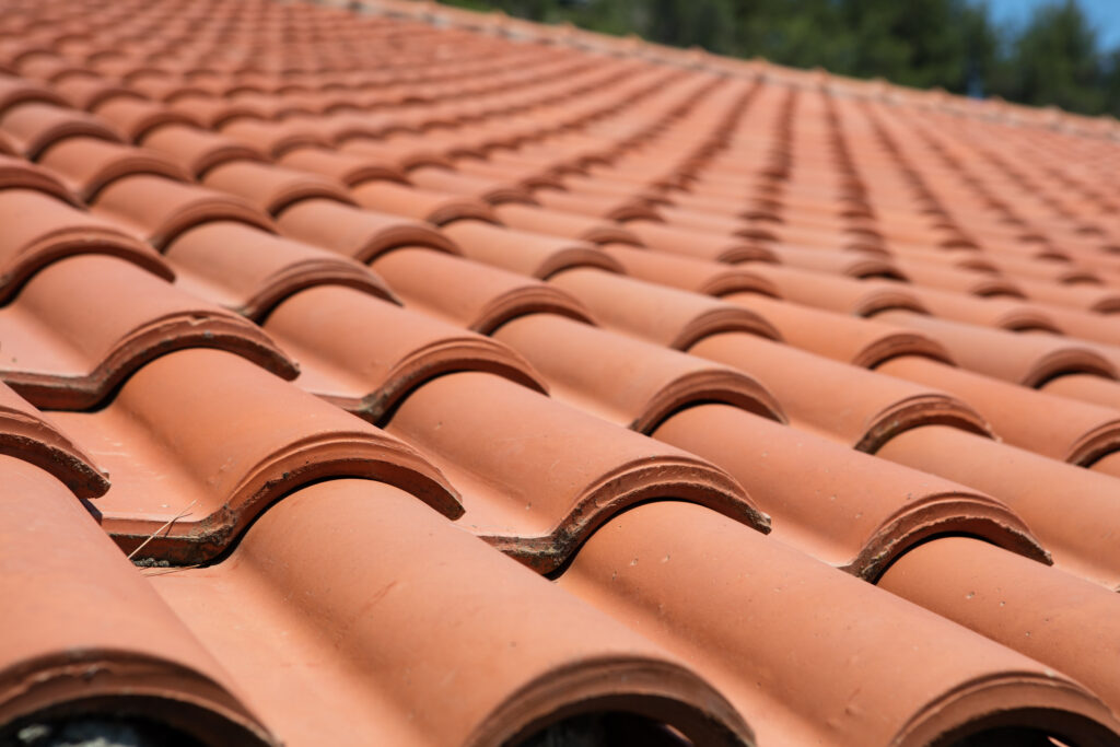 Best Roofing Materials Alexandria VA Close-up view of a roof covered with overlapping terracotta clay tiles, arranged in neat, horizontal rows. The tiles create a wavy pattern, and trees are visible blurred in the background.
