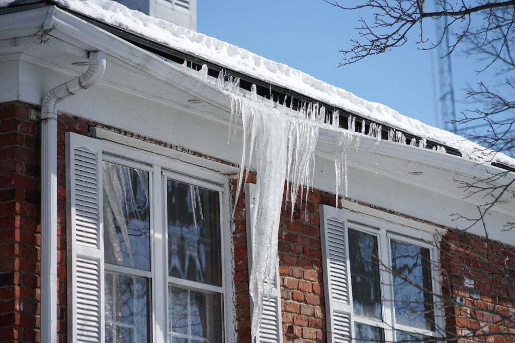 winter roof replacement Virginia Icicles hang from the snow-covered edge of a roof on a brick house with white shutters and double windows, with bare tree branches in the foreground.