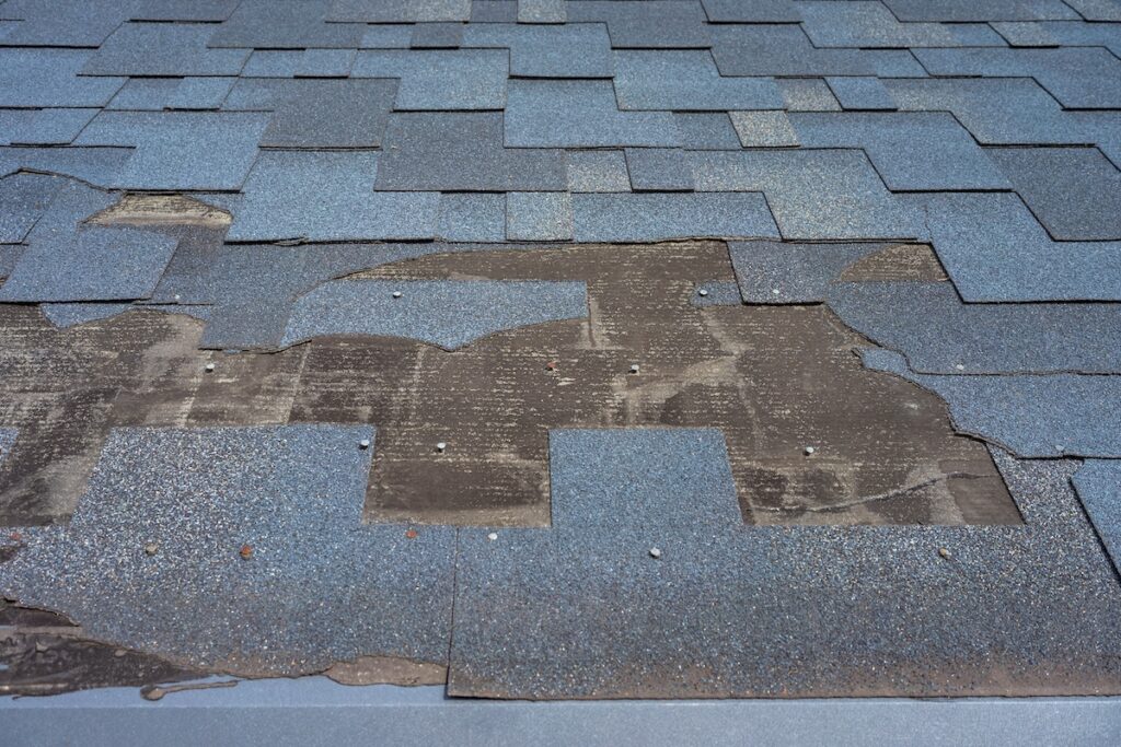 Emergency Roof Repair Alexandria VA A section of a blue shingle roof with missing and damaged shingles, exposing the underlayment and nails beneath. The area shows significant wear and needs repair.