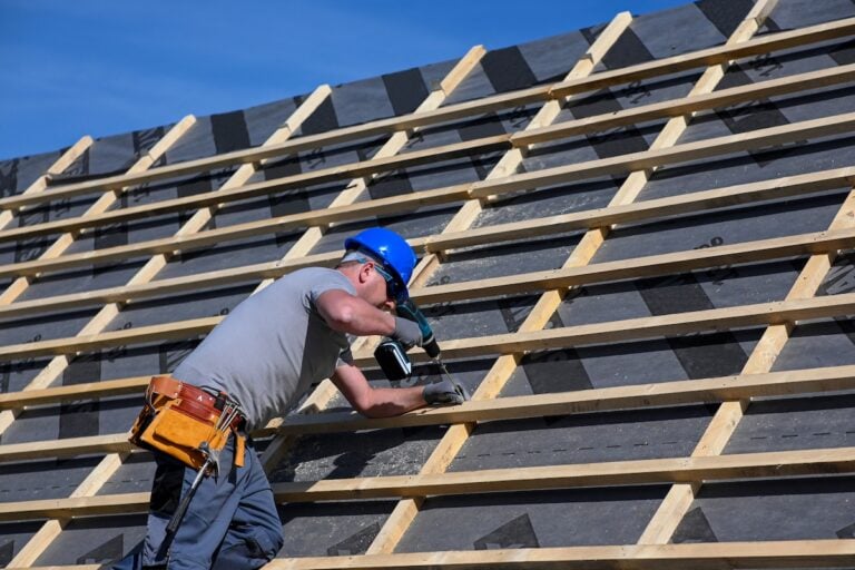 A construction worker in a blue hard hat uses a power drill to secure wooden beams on a slanted roof under clear blue sky. The worker wears a tool belt and safety gloves.