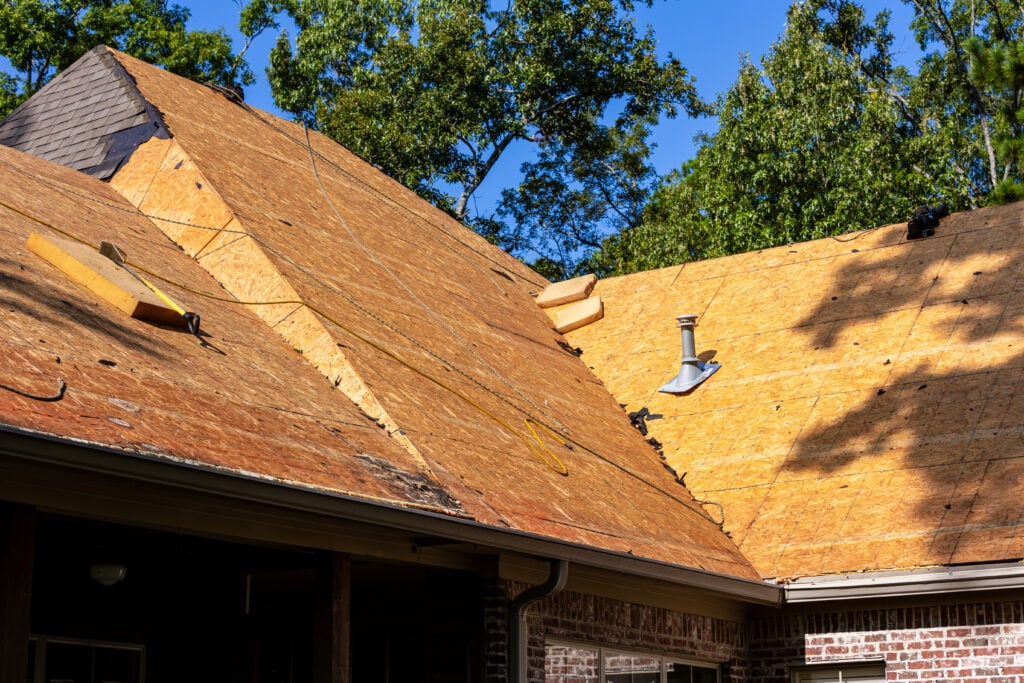 Roof Replacement Cost Alexandria VA Partially constructed house roof with plywood sheathing, roof vents, and tools visible; surrounded by trees under a clear blue sky.