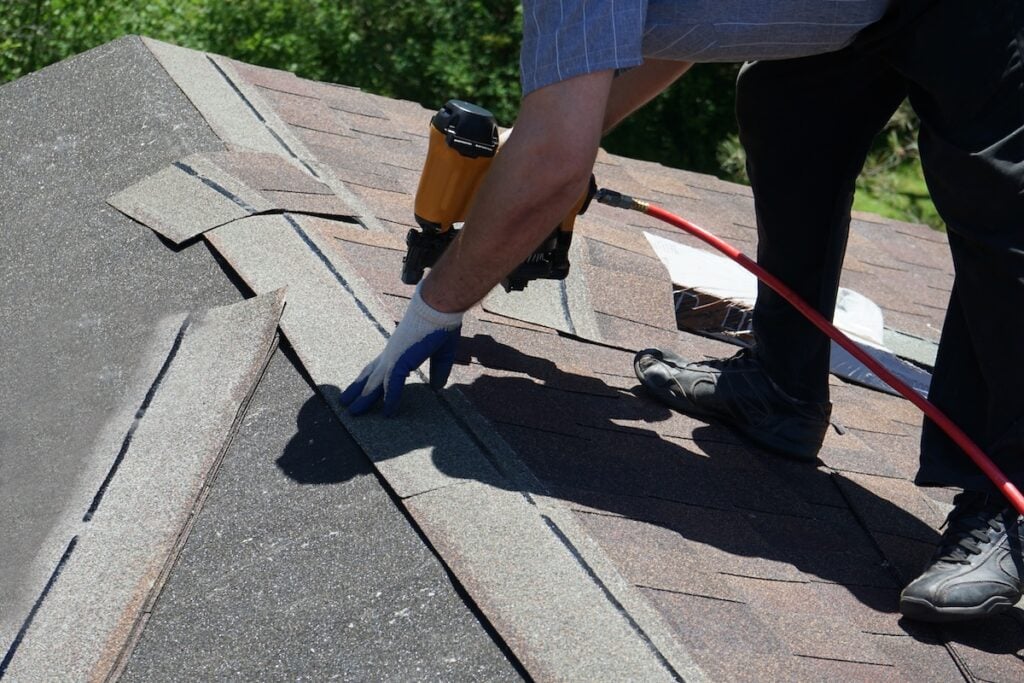Roof Replacement Cost Alexandria VA A person wearing gloves uses a nail gun to install asphalt shingles on a roof, working at the peak where two roof sections meet. Tools and materials are visible nearby.