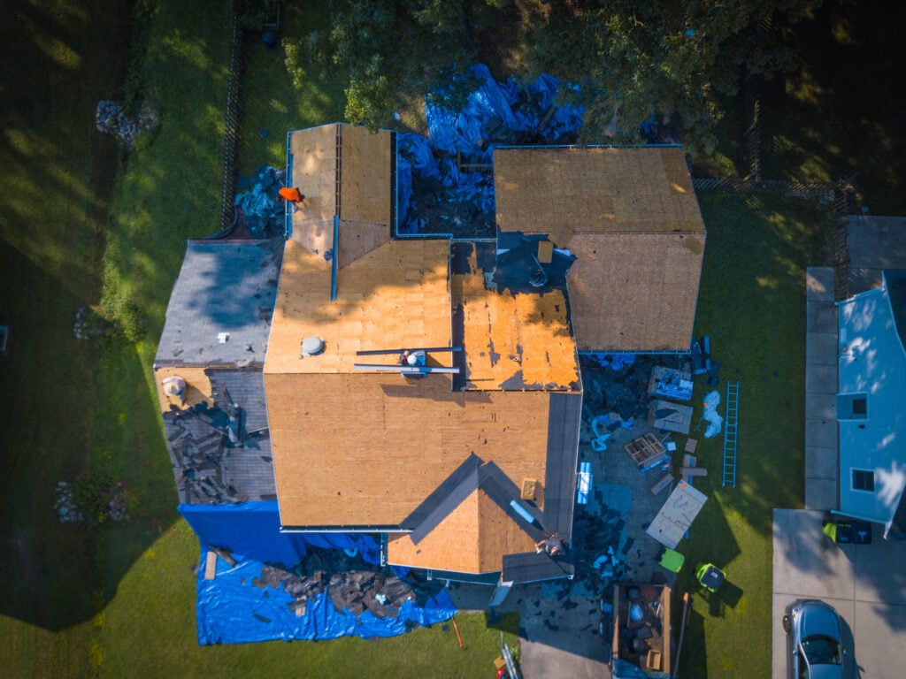 Roofing Permits Alexandria VA Aerial view of a house with workers repairing or replacing the roof. Sections of the roof are covered with tarps and exposed plywood. Tools, materials, and debris are scattered around the yard below.