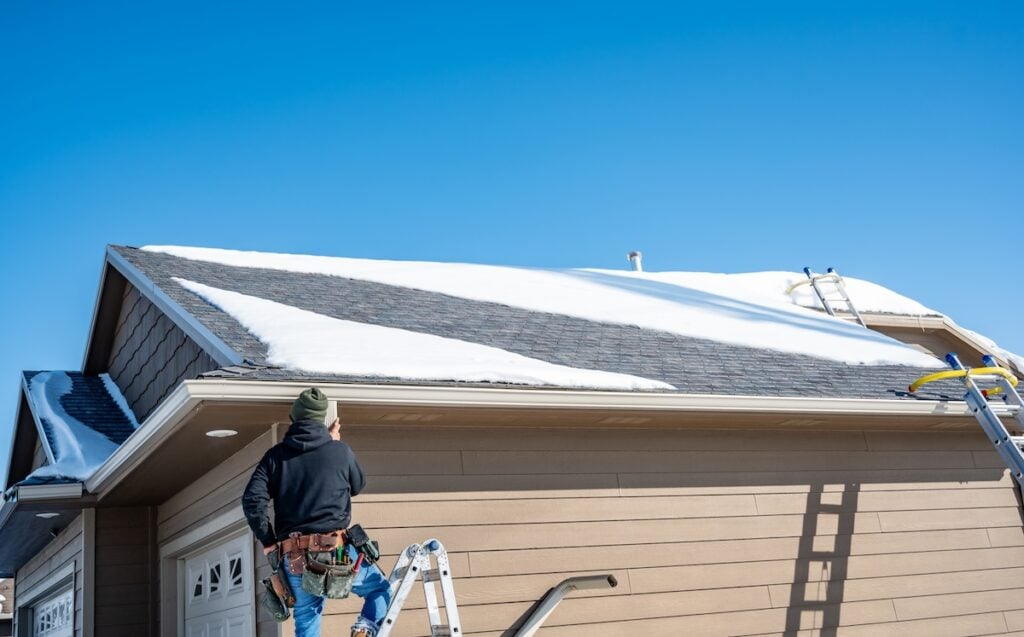 winter roof replacement Virginia A person in winter clothing stands on a ladder next to a house, looking up at a snow-covered roof under a clear blue sky. Another ladder and roof rake are positioned against the roof.