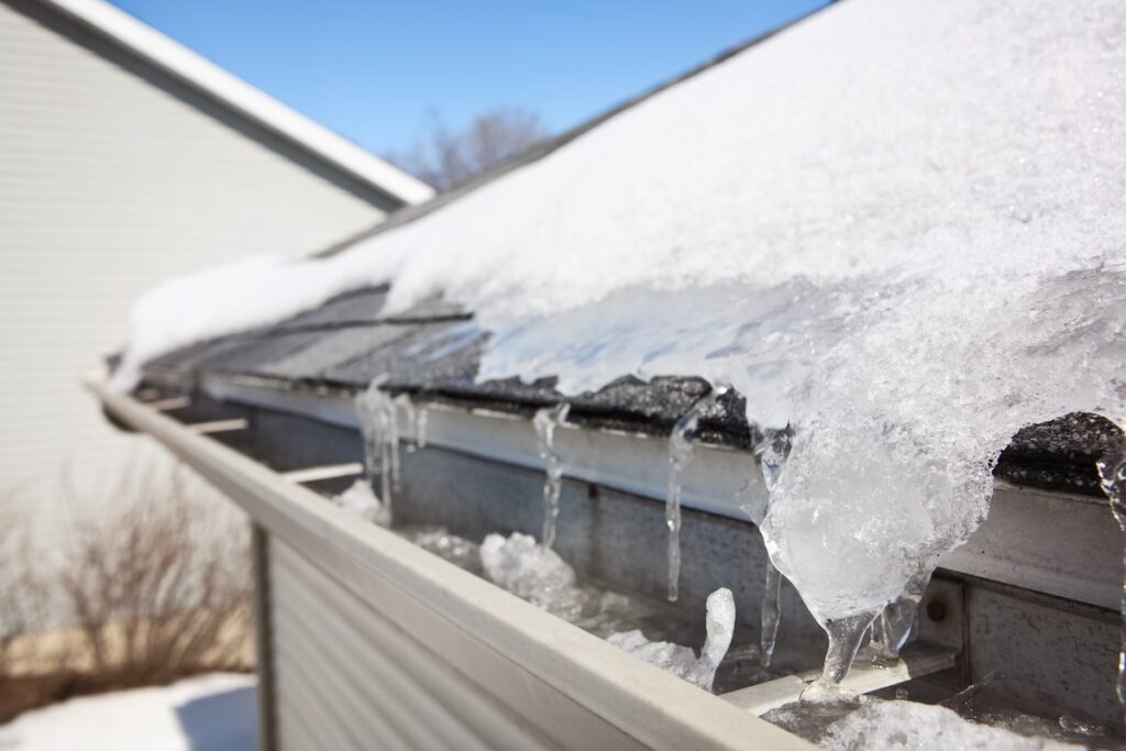 Roof Damage McLean Close-up of a house roof covered in snow and ice, icicles hanging from the edge and gutter—a common cause of roof damage in McLean. Clear blue sky above, with bare ground and leafless shrubs visible below.