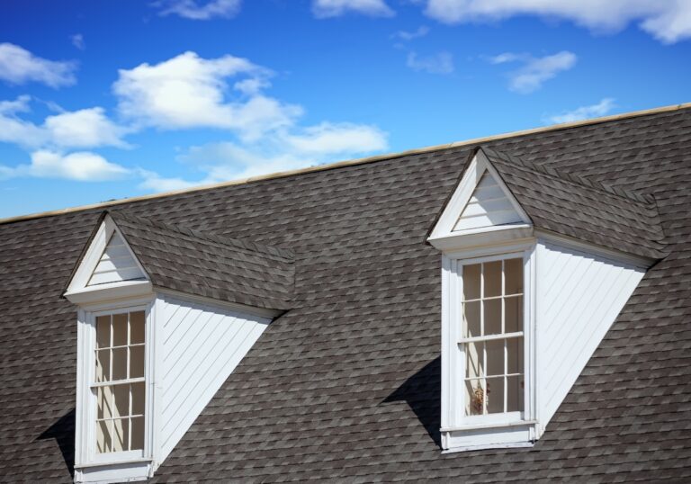 Two white dormer windows with triangular roofs are set into a gray shingled roof under a bright blue sky with scattered white clouds.