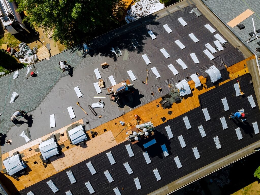 Aerial view of workers installing new roofing materials on a large building; bundles of shingles, tools, and equipment are spread across the roof, with partial sections covered in underlayment.
