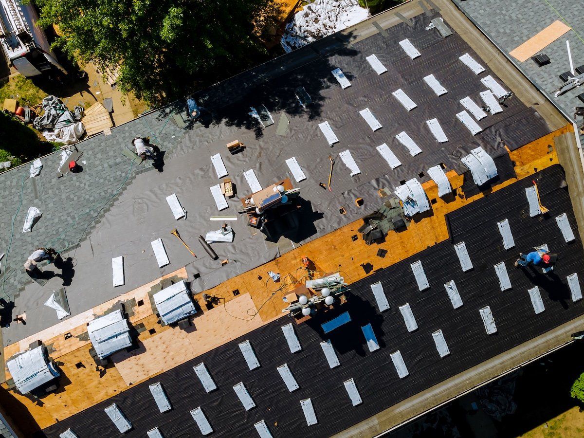 Aerial view of workers installing new roofing materials on a large building; bundles of shingles, tools, and equipment are spread across the roof, with partial sections covered in underlayment.
