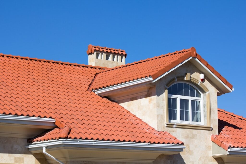 Energy Efficient Roofing Virginia A close-up view of a house with beige stone walls and a red tiled roof, featuring a chimney and an arched window, set against a clear blue sky.